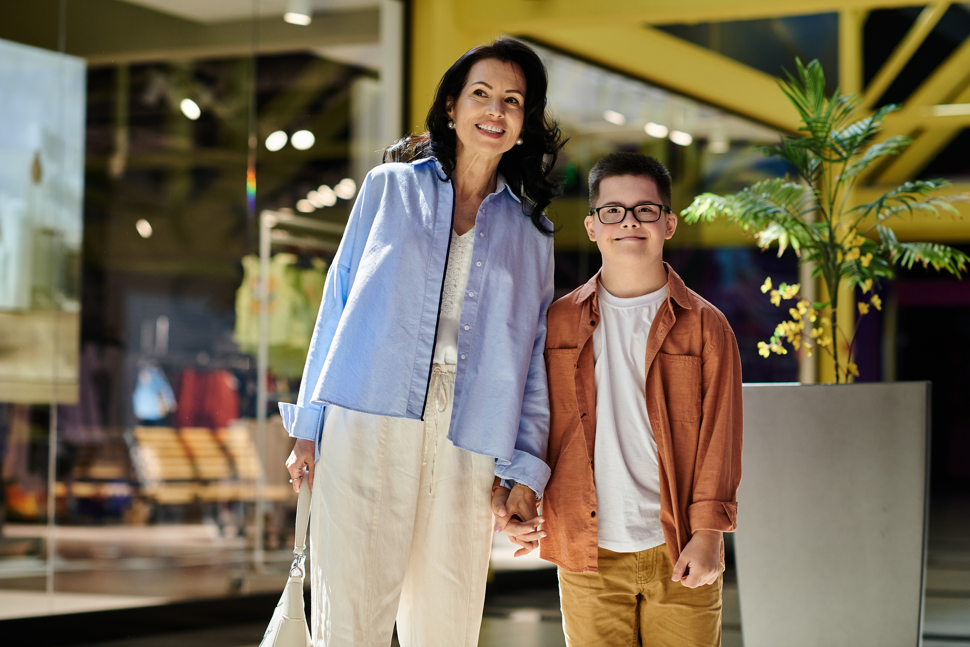 A mother and her son with Down syndrome enjoy a day out together, walking through a brightly lit shopping mall.